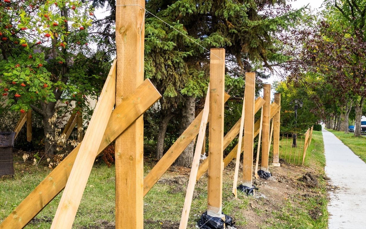 A wooden fence mid-installation in a yard near trees and a sidewalk.