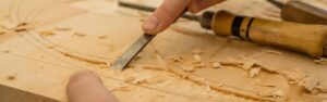 Hands use a chisel to carve a decorative pattern into a flat piece of light-colored wood.
