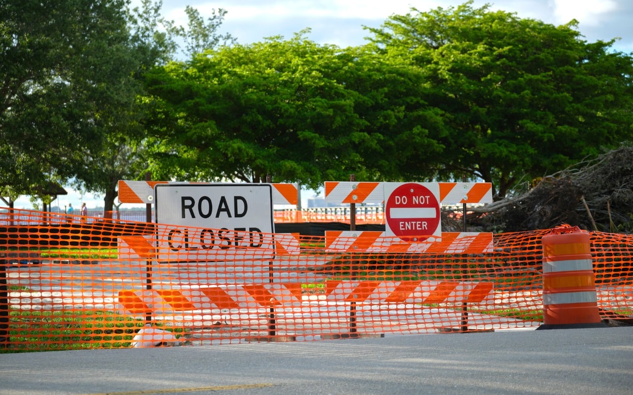 Orange safety fencing and road closure signs protect mature trees in the background during a construction project.