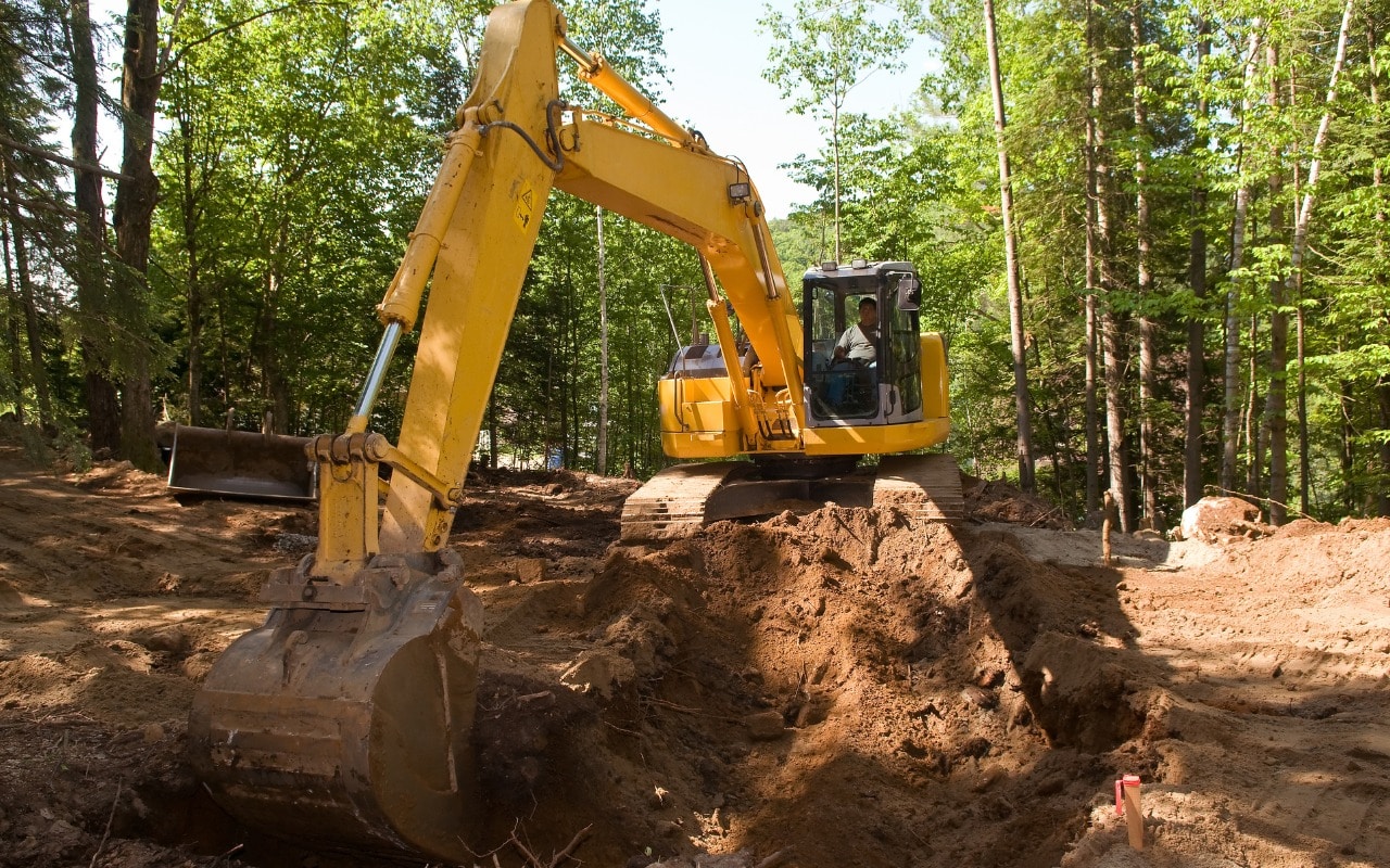 An aerial view shows an excavator digging into red clay soil near a wooded area with visible trees in the background.