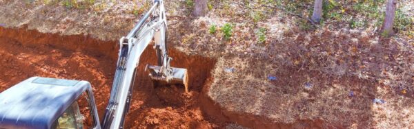 A yellow excavator with a bucket attachment digs into brown soil at a wooded construction site surrounded by mature trees.