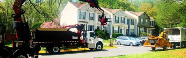 A truck-mounted crane is set up on a residential street with its boom raised beside two-story homes and mature trees.