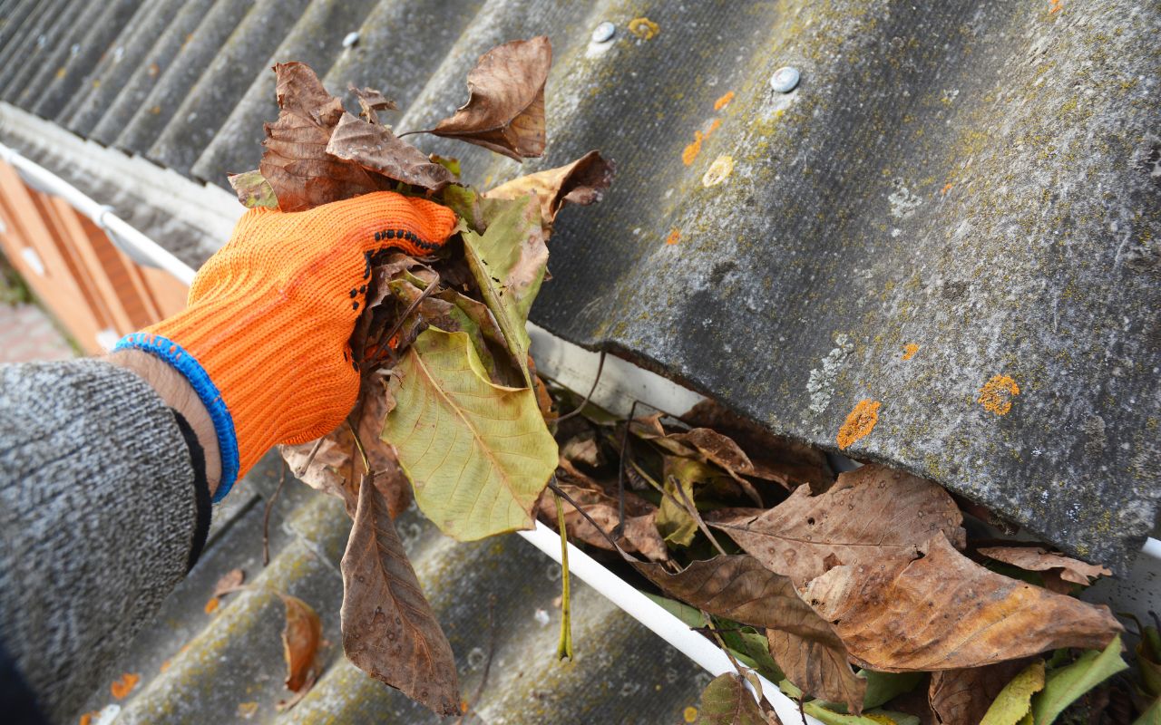 A person with orange gloves pulls dried leaves out of a clogged gutter.